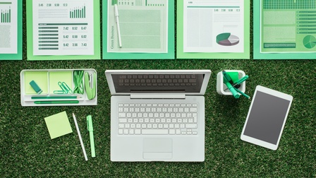 Vogelperspektive auf aufgeklapptem Laptop auf Rasen platziert, umgeben von Dokumenten und Büroutensilien in grün