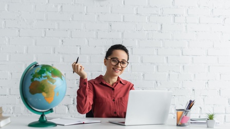 Person mit geschlossenen dunklen Haaren und Brille sowie roter Bluse sitzt bei einem Schreibtisch und blickt freudig darauf während ein Stift in der Hand gehalten wird, am Tisch steht ein Globus, ein Stiftebecher sowie weitere Unterlagen liegen darauf