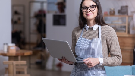 Person mit Brillen in Schürze lehnt an Tisch und hält aufgeklapptes Notebook in Händen, im Hintergrund verschwommen Gästebereich eines Kaffeehauses