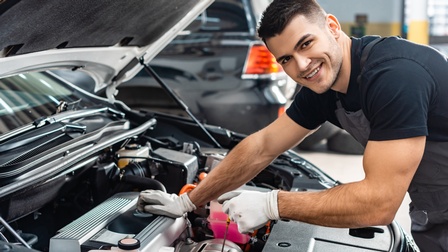 Seitliche Porträtaufnahme einer lächelnden Person mit Overall und Handschuhen, die neben einer geöffneten Motorhaube steht und ein Stäbchen zur Messung des Ölstandes hält