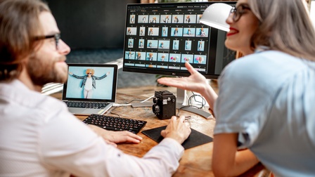 Zwei Personen unterhalten sich bei einem Schreibtisch, darauf stehen ein Laptop sowie ein Monitor mit ersichtlichen Fotos sowie steht eine alte Rollei Kamera auf dem Tisch
