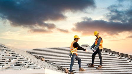 Zwei Personen mit Helmen, Sicherheitswesten und Handschuhen stehen auf einem Dach eines Gebäudes. Sie halten gemeinsam eine graue, flache Fliese. Links neben den Personen sind weitere gestapelte Fliesen. Der Himmel ist bewölkt