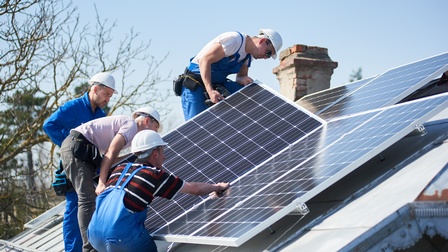 Vier Personen mit blauen Arbeitsoveralls und weißen Helmen auf Dach stehend bei der Montage und Inspektion von Solarpanelen