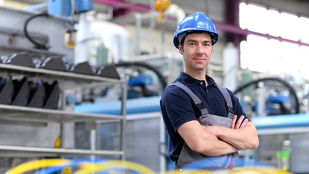 Portrait einer Person mit Schutzhelm und Arbeitshose. Sie hat die Arme vor dem Körper verschränkt. Im Hintergrund ist eine Industrieanlage zu sehen.