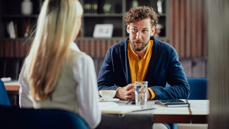 Zwei Personen sitzen sich an einem Tisch gegenüber. Die der Kamera zugewandte Person hat einen Bart und braune, kurze Haare. Auf dem Tisch sind ein Tablet und eine Tasse Kaffee. Im Hintergrund sind weitere Tische