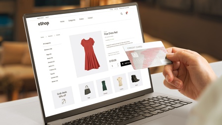 Eine Person kauft online ein rotes Kleid und hält dabei ihre Kreditkarte vor den Laptop, bereit zum Bezahlen.