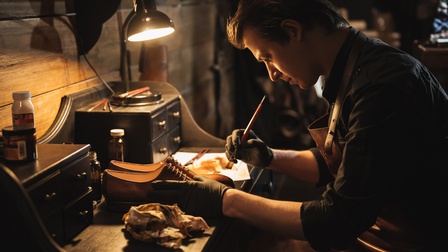Person sitzt über beleuchtete Werkbank gebeugt und bearbeitet Lederschuh,, ringsum Arbeitsutensilien