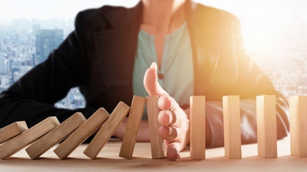 Die Büste einer Person ist zu sehen, sie sitzt an einem Holztisch. Mit der Hand stößt sie rechteckige Bausteine um. Im Hintergrund sieht man Umrisse einer Großstadt mit Hochhäusern.