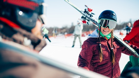 Nahaufnahme von zwei Personen, eine von vorne, eine von hinten. Sie stehen sich gegenüber und halten Skier in der Hand, haben Skihelme und Skibrillen auf. Im Hintergrund ist eine Skipiste mit weiteren Personen 
