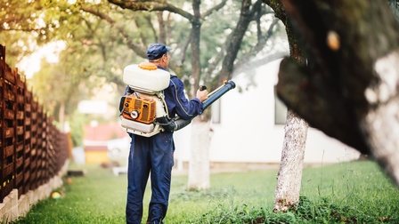 Person in Rückansicht mit Container und Minimotor am Rücken steht in Wiese zwischen Holzzaun und Bäumen und hält Röhre, die mit Container verbunden ist, in die Luft, im Hintergrund verschwommen weitere Bäume und Hausfront