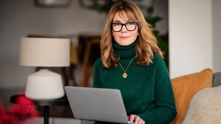 Porträt einer Person mit Brillen in grünem Wollkragenpullover mit aufgeklapptem Laptop am Schoß, im Hintergrund verschwommen Wohnraum