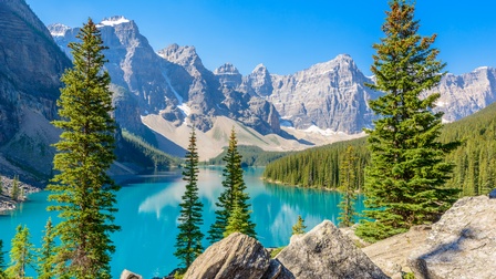 Schroffe Bergkette umsäumt von Nadelwäldern und türkisblauem Gebirgssee, im Vordergrund einzelne Bäume und Felsen