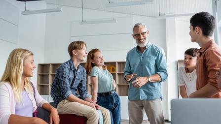 Mehrere Jugendliche in Klassenraum stehend und lachend, in der Personengruppe mittig Erwachsener