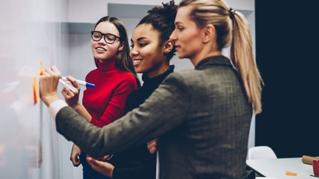 Seitliche Aufnahme von drei Personen, die nebeneinander an einem Whiteboard stehen. Zwei der Personen notieren jeweils etwas auf einem Post-it, die auf dem Whiteboard kleben