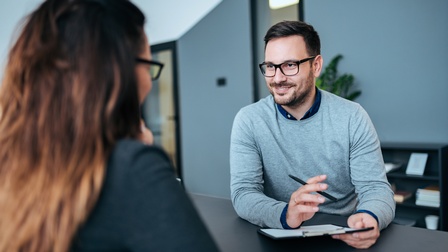 Person mit dunklen kurzen Haaren, Brille und Bart sitzt an einem Tisch und hält ein Klemmbrett sowie ein Stift in Händen während mit einer anderen Person gesprochen wird