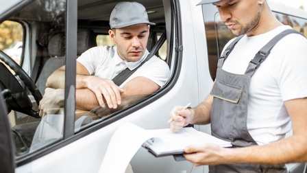 Am Steuer eines geparkten Kleinlastwagens sitzt eine Person mit einer Kappe. Neben ihr steht eine weitere Person mit Kappe, die ein Klemmbrett hält. Beide Personen blicken auf das Klemmbrett