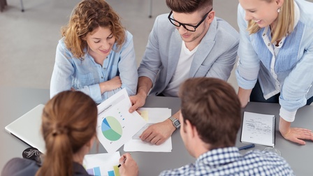 Eine Gruppe aus Personen sitzt und steht rund um einen Tisch und blickt auf verschiedene Unterlagen, Vogelperspektive