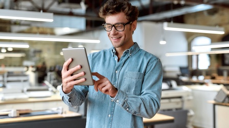 Eine Person mit Brille, Bart und Jeanshemd hält ein Tablet in den Händen, worauf sie auch blickt und mit der linken Hand klickt. Im Hintergrund sind Tische und von der Decke hängende Lampen