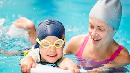 Kind und erwachsene Person in Wasser badend mit Gummihauben, Kind trägt gelbe Schwimmbrille und hält sich an Schwimmbrett fest, erwachsene Person stützt Kind beim Schwimmen