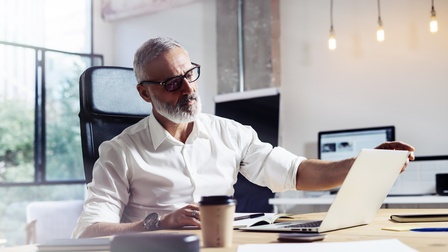 Person mit kurzen grauen Haaren, Bart, Brille und weißem Hemd sitzt bei einem Schreibtisch in einem Büro mit aufgeklappten Laptop, weitere Unterlagen liegen auf dem Tisch 