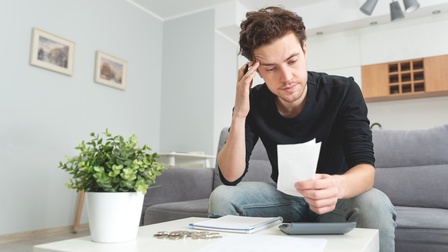 Eine Person sitzt auf einem Sofa. Vor ihr ist ein niedriger, heller Tisch. In der linken Hand hält die Person kleine Zettel, auf die sie blickt. In der rechten Hand hält sie einen Stift. Auf dem Tisch sind ein Taschenrechner, ein Notizheft und Münzen