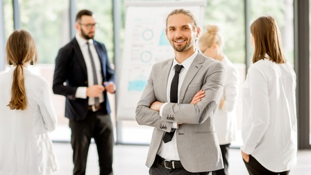 Portrait einer Person mit geschlossenen Haaren und Bart in Businesskleidung, im Hintergrund stehen weitere Personen rund um ein Flipchart