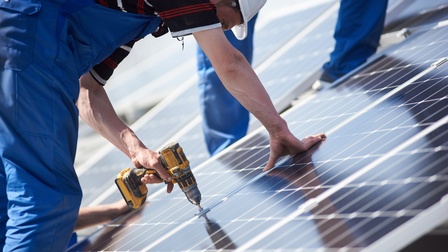 Person in blauer Arbeitslatzhose mit weißem Helm mit Bohrmaschine in der Hand beim der Montage von Solarpanelen