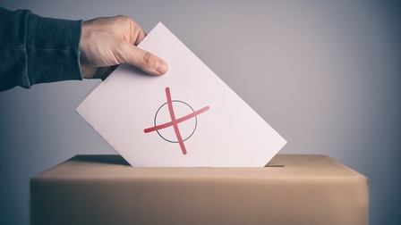 Seitliche Aufnahme einer Wahlurne, in die eine Person einen weißen Zettel wirft, auf dem ein schwarzer Kreis ist, in dem ein rotes X ist