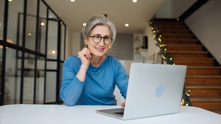 Lächelnde Person mit Brillen an Schreibtisch vor aufgeklappten Laptop sitzend und darauf blickten, im Hintergrund verschwommen Holztreppe mit Pflanzengirlande mit Leuchtkette am Geländer