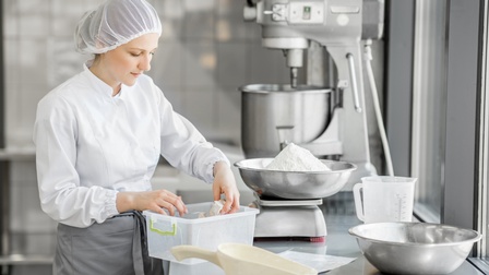 Person mit weißem Haarnetz und weißer Arbeitsjacke sowie grauer Schürze bereitet Zutaten für Backwaren vor