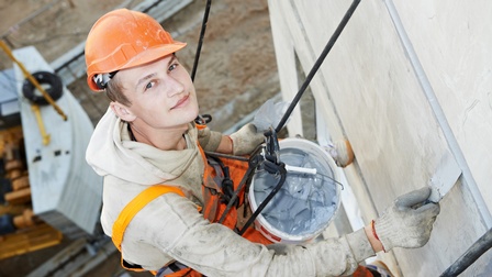 Person mit oranger Schutzkleidung sowie Schutzhelm blickt in die Kamera, hängt mit Sicherung an Hausfassade und bearbeitet diese