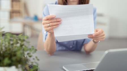 Nahaufnahme einer Person mit braunen Haaren, die konzentriert auf ein Dokument aus Papier blickt, das sie in beiden Händen hält. Sie steht und hat die Arme auf einen grauen Tisch gestützt, auf dem ein aufgeklappter Laptop und eine Topfpflanze steht