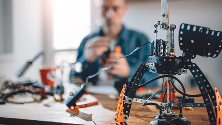 Ein technisches Werkstück ist im Fokus während dahinter in der Unschärfe eine Person an etwas arbeitet