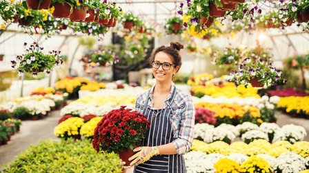 Lächelnde Person mit Arbeitsschürze und Handschuhen hält Blumen in einer großen Gewächshalle mit weiteren Blumen und Pflanzen 