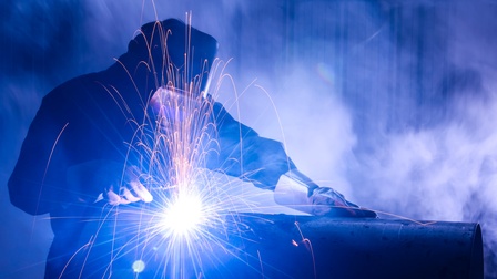 Person mit Schweißhelm und dicken Arbeitshandschuhen beugt sich über ein dickes Metallrohr, um mit einem Schweißgerät dieses zu bearbeiten. Daneben Funken und einen sehr hellen Punkt in der Bildmitte, wo geschweißt wird