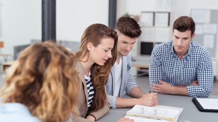 An einem Tisch sitzen vier Person. Auf dem Tisch liegen Unterlagen sowie ein aufgeklappter, dicker Ordner mit vielen Zetteln. Die Personen blicken auf die Zettel im Ordner