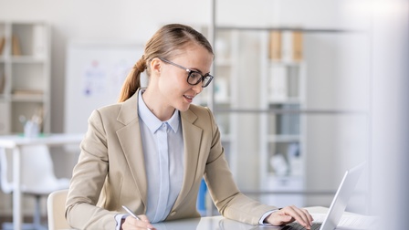 Person in Businesskleidung, geschlossenen Haaren und Brille sitzt an einem Schreibtisch, blickt auf einen Laptopbildschirm und macht sich Notizen