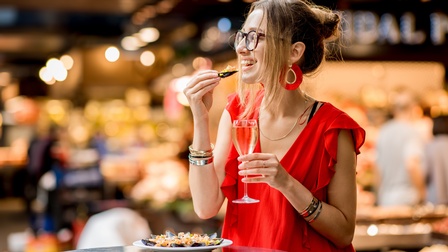 Person mit dunklen geschlossenen Haaren, Brille und rotem Shirt isst genüsslich Muscheln und trinkt Sekt bei einem Stehtisch