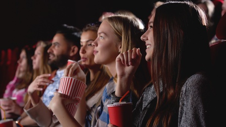 Mehrere Personen nebeneinander in Kinosaal sitzend schauen Richtung Leinwand und halten Getränke und Popcorn in Händen