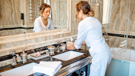 Rückenansicht einer Person in heller Uniform, die in einem Hotelzimmer im Badezimmer steht und die Hände unter den Wasserhahn hält