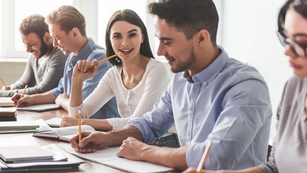 Seitliche Aufnahme von fünf Jugendlichen, die nebeneinander an einem Tisch sitzen und etwas in Heften notieren. Eine der Personen blickt direkt in die Kamera