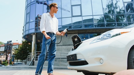 Eine Person hält ein Ladekabel bereit, um ein weißes Elektroauto aufzuladen. Im Hintergrund befindet sich ein Gebäude mit Glasfassade.