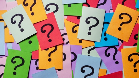 Topshot vieler kreuz und quer sowie teilweise übereinander liegender, bunter Zettel. Auf jedem Zettel ist ein schwarzes Fragezeichen
