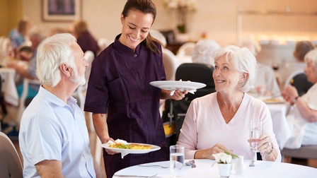 Zwei Personen sitzen in einem Restaurant an einem Tisch. Neben ihnen steht eine Person, die in jeder Hand jeweils einen Teller mit einer Speise hält und diese vor den Leuten abstellt. Im Hintergrund sitzen viele weitere Personen an Tischen