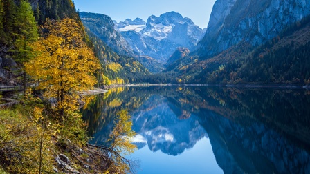 Schroffe Bergkette unter blauem Himmel umfriedet von grünen Wiesen, im Vordergrund Bergsee, in dem sich die Bergkette spiegelt
