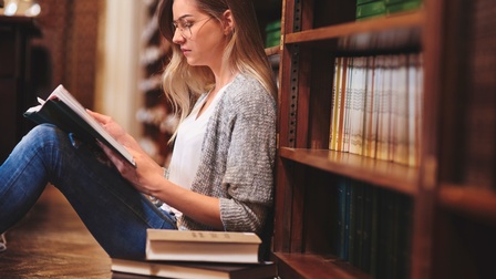Person mit Brillen sitzt am Boden einer Bibliothek und liest in aufgeschlagenem Buch