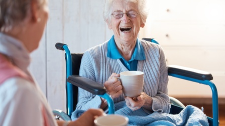 Lachende ältere Person mit Brillen sitzt in Rollstuhl und hält Tasse in der Hand, im Vordergrund verschwommen weitere Person mit Tasse