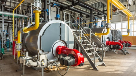 Drei große graue Kesselanlagen umgeben und verbunden mit zahlreichen gelben, schwarzen, roten und chromfarbenen Verrohrungen in Industriehalle