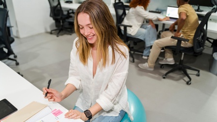Eine jugendliche Person mit offenen, hellen Haaren sitzt auf einem Sportball an einem Schreibtisch in einem Raum mit mehreren Schreibtischen. Vor ihr ist ein aufgeklapptes Notizbuch, in dem verschiedene Post-its kleben. Die Person hält einen Stift