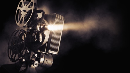 Ein Filmprojektor vor schwarzem Hintergrund, von dem ein heller Lichtschein ausgeht.